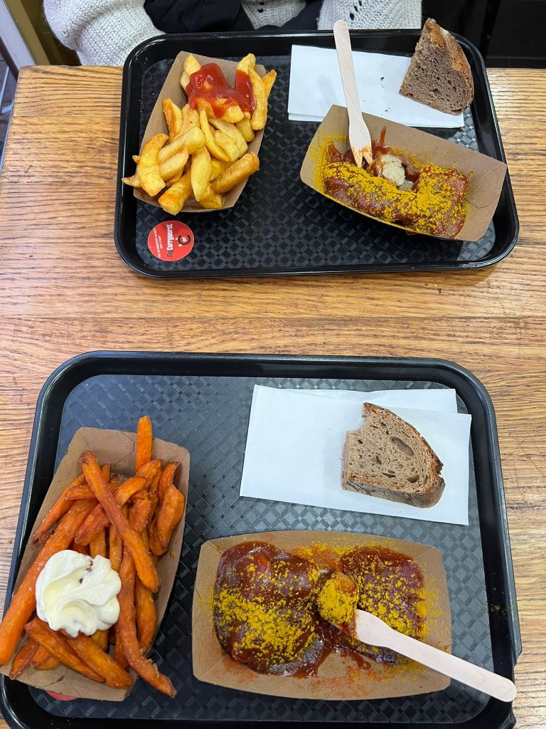 Photo showing two meals on plastic trays and wooden table. Both have a piece of bread and a tray with sausage covered in a red paste and curry powder. One meal has sweet potato fries and mayo on the side while the other has normal fries with ketchup.