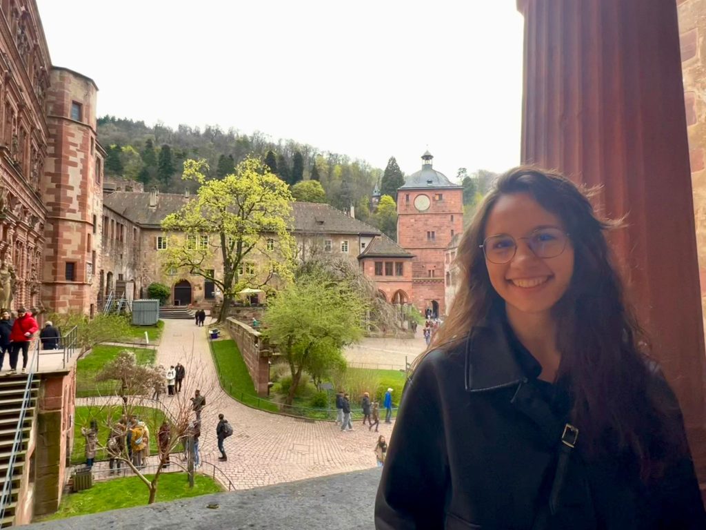 A brunette girl smiling with a view of a castle courtyard in the background. The courtyard has green trees, paved paths with tourists walking and in surrounded by old red brick buildings.