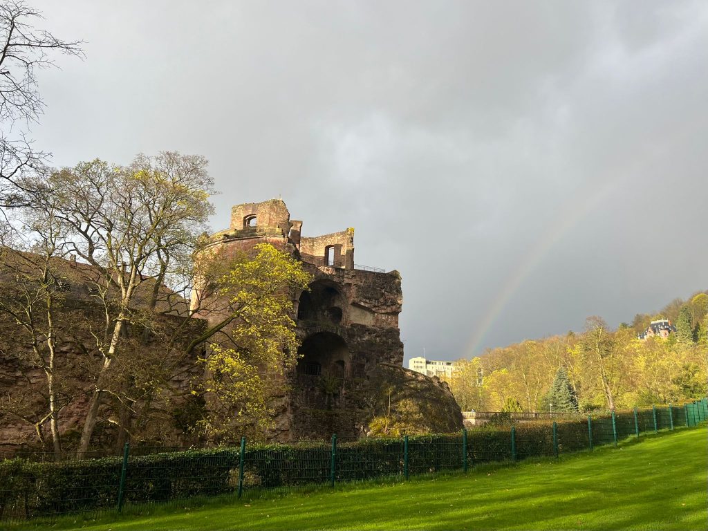 Photo of the ruins of the Heidelberg castle. It is made out of red brick and surrounded by greenery. In the background, a cloudy sky and a rainbow can be seen.