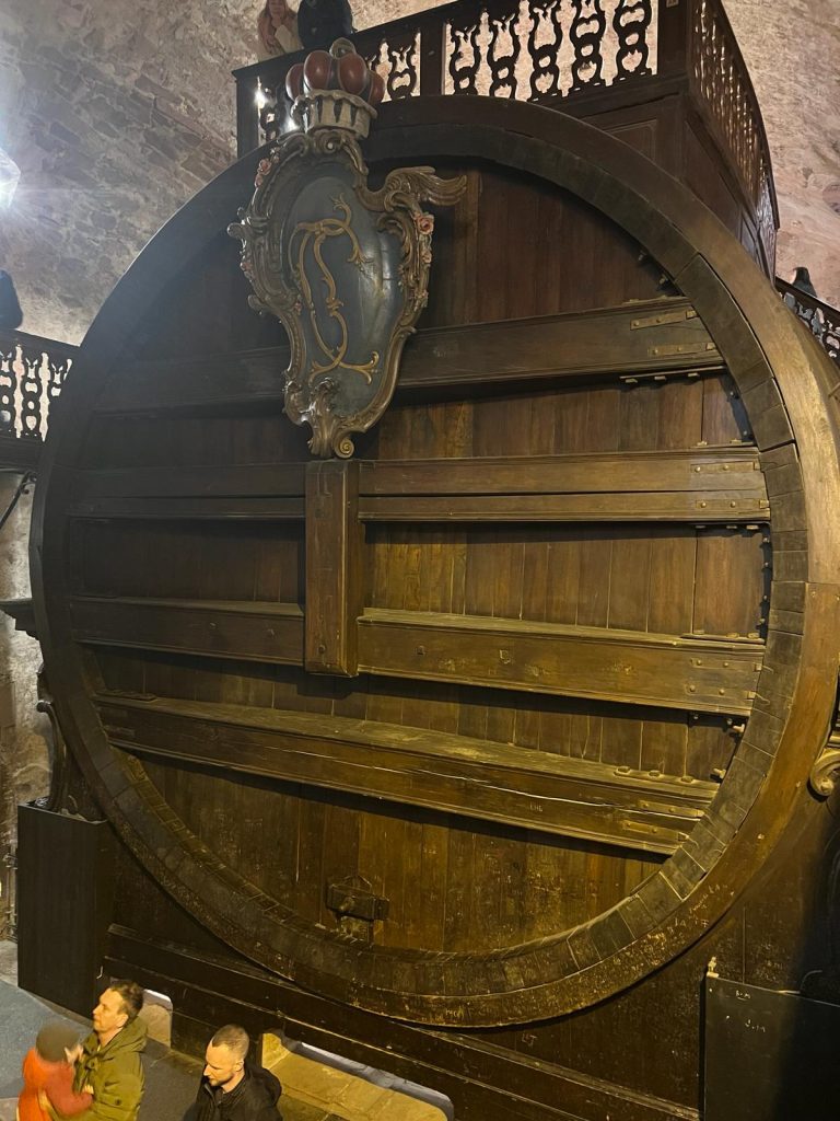 Photo of an insanely large wine barrel. It is made out of wood and has a coat sculpture at the top. a few people can be seen around it.