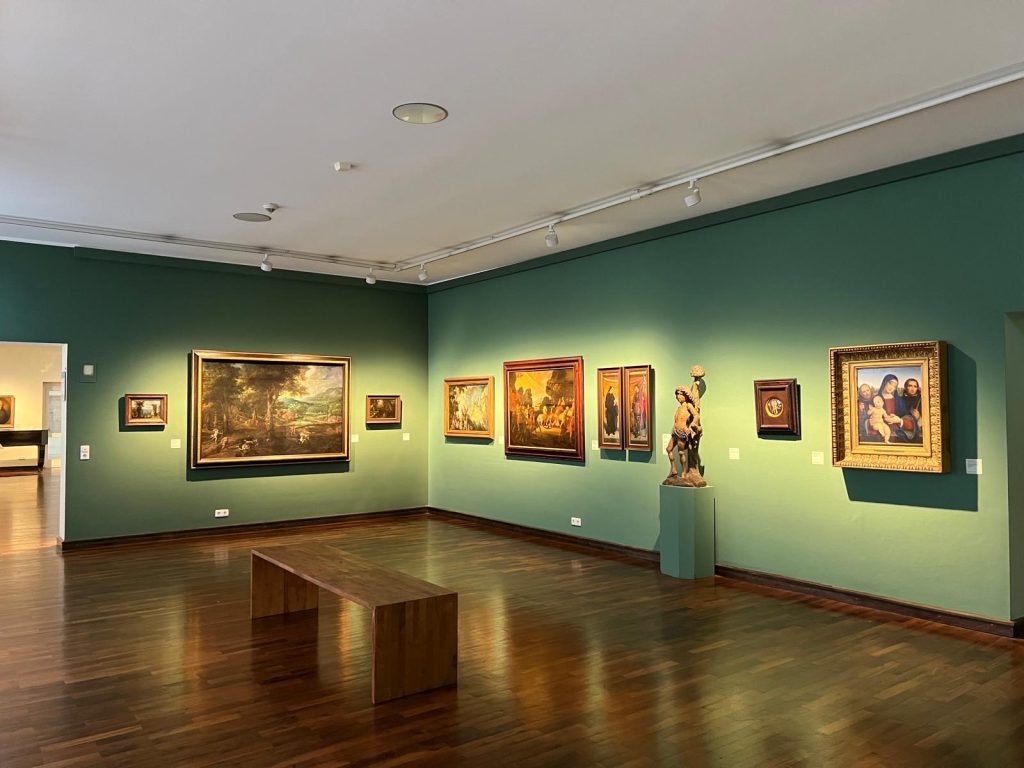 Photo of the inside of an art useum. The walls are light green and numerous old paintings are hung up. There is also a small sculpture and the floor is wooden.