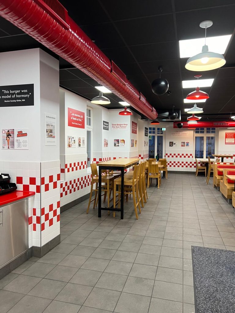 Inside a American style restaurant. The walls are covered in red and white tile, wooden chairs and tables can be seen and the floor is gray.