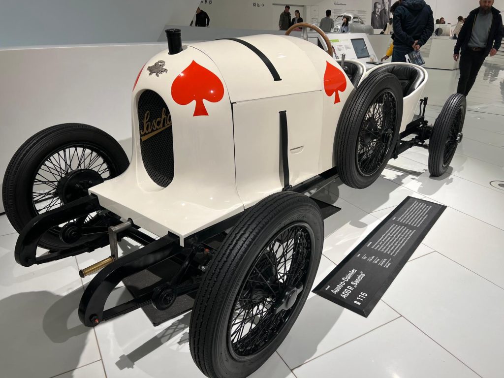 Photo of a vintage Porsche car model. The car is white and has two  red ace symbols painted on it. The wheels are thin and black.