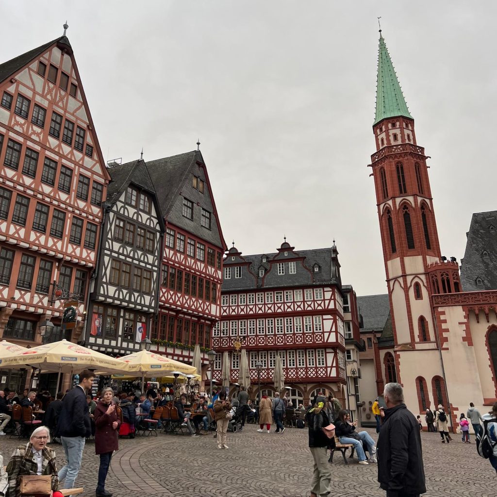 Image of a old square in Frankfurt. Traditional German building are seen on the left and a church tower can be seen on the right. People are sitting down at tables.