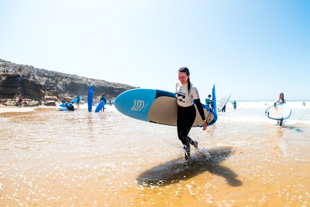 Annmari går på stranden upp från havet och håller i en surfbräda.