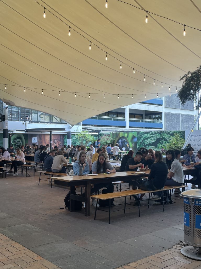 students sitting at long tables under the fairy lights and tarp at the quad of UoA
