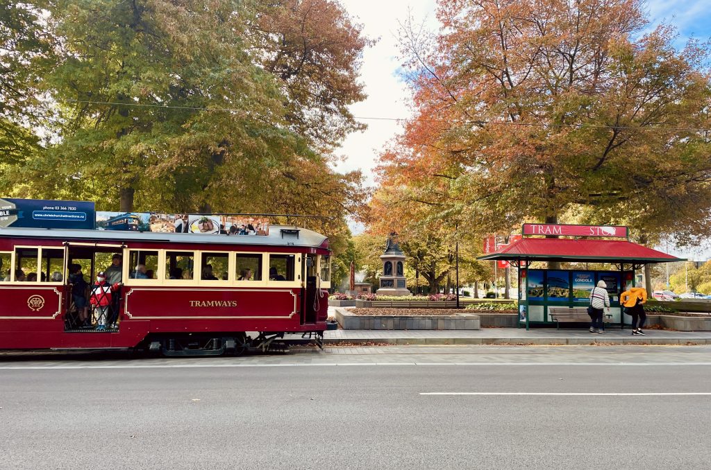 old-fashioned tram approaching a bus stop near an autumn-coloured park in Christchurch