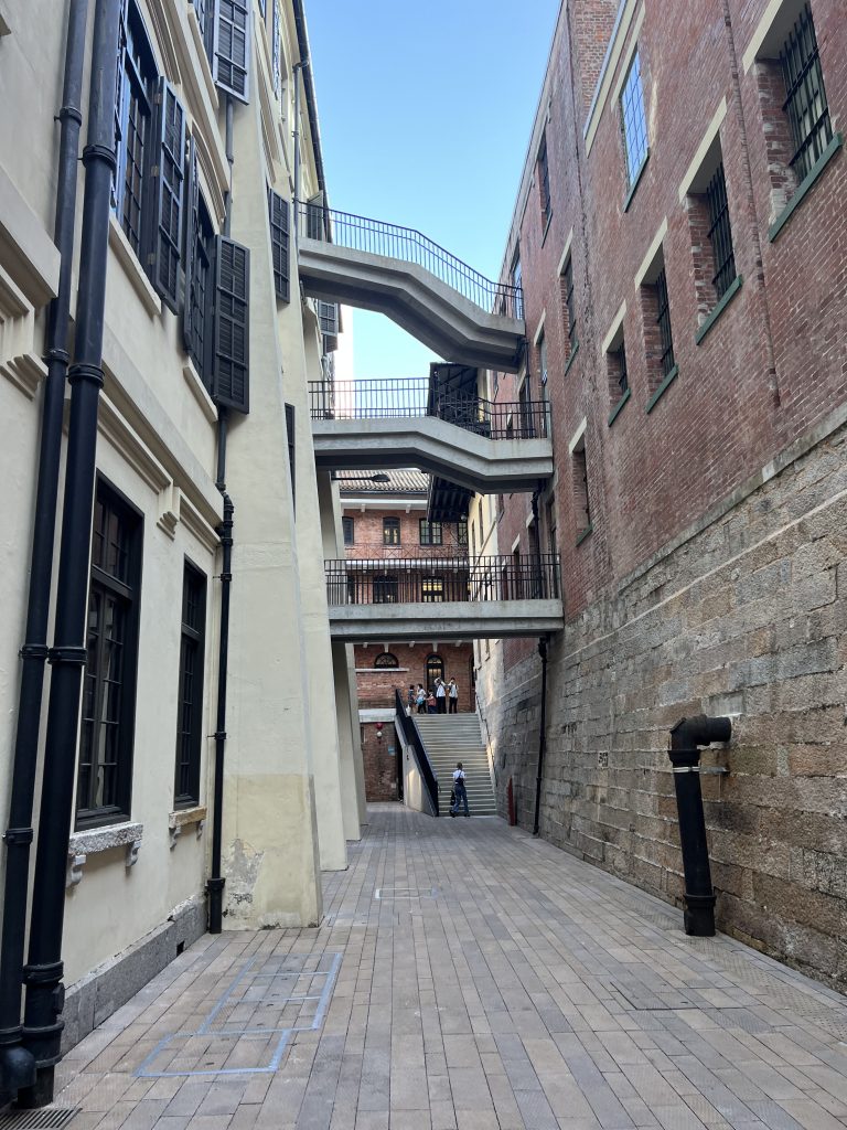 Two buildings connected by three bridges at different levels. One building is white with black windows and is the old polisstation. The other building is made of red tiles and is the old prison of Tai Kwun in Hong Kong.