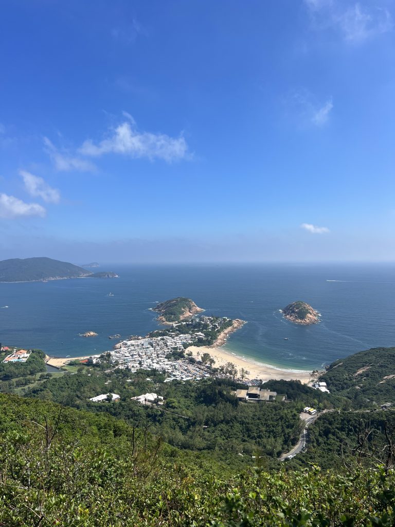 A picture taken from the top of a mountain overlooking a city of white buildings. Leading up to the city is greenery and behind it is the blue oceans and sky with several smaller islands interspersed. 