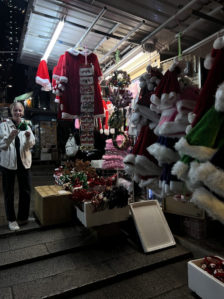 A girl standing infront of stalls selling christmas items such as santa outfits and head bands with christmas trees on