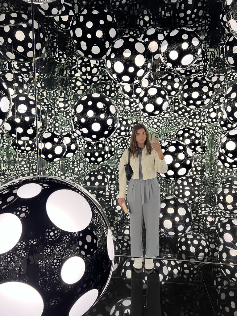 Girl standing and taking a photo in a room full of black and white balls and mirror, looking like abstract art at the M+ museum