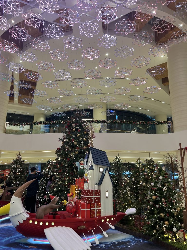 Shopping mall christmas decorations with many small christmas trees as well as an otter and fairy on a flying boat with presents.