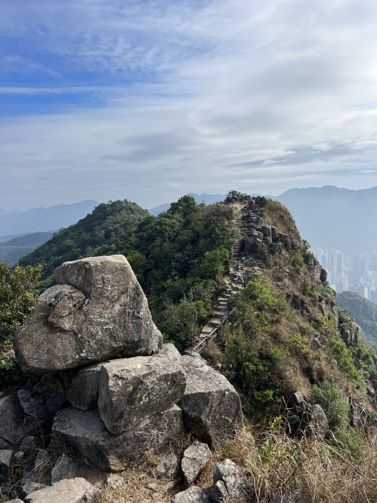 A mountain peak with a stone path leading up to the top. Surrounded by greenery and in the background you can see the faint outline of skyscrapers and other mountains.
