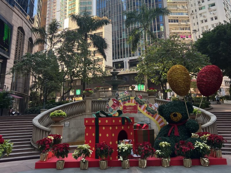 Christmas decorations in the city, in the area of Sheung Wan. A teddy bar holding two balloons next to two presents and flowers. The decorations are infront of two stair cases, several trees and sky scrapers in the background.