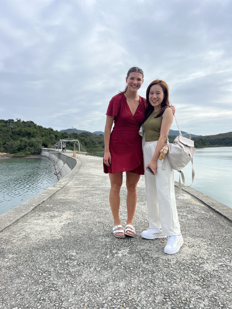 Two girls standing on a stone bridge and smiling for the camera surrounded by water and leading to an island with greenery in the distance.