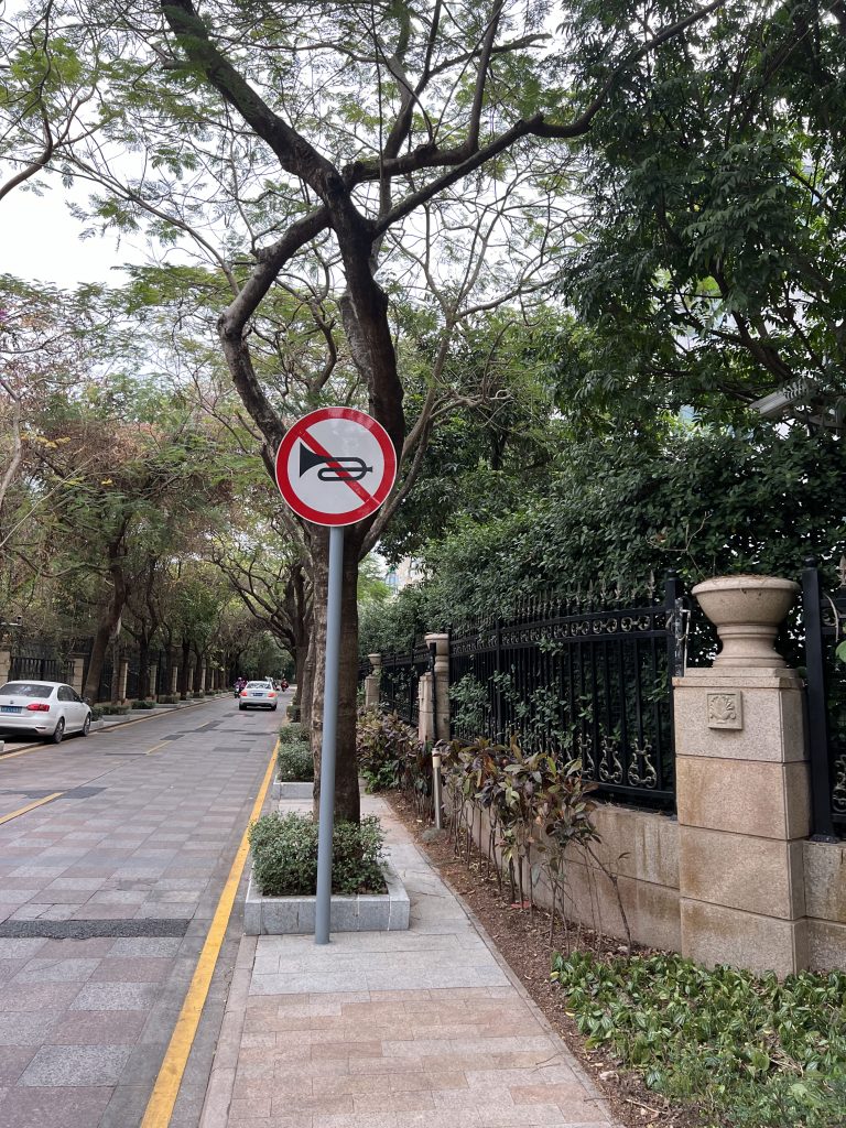 A sign indicating no honking by showing a trumpet crossed over on a street with trees. 