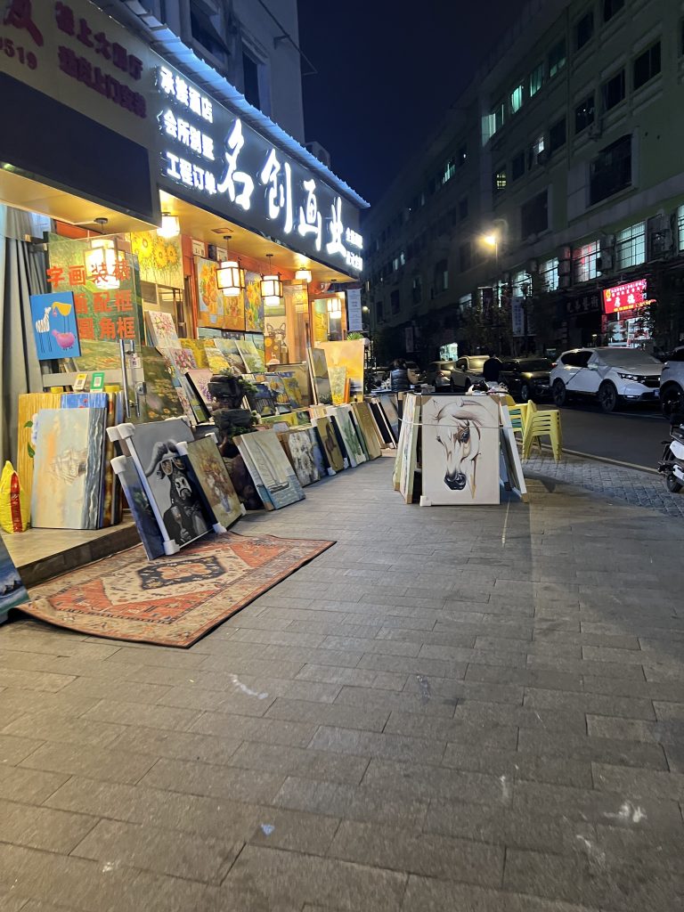 A corner shop selling oil paintings at night time. The paintings are leaning on each other and the motifs include a horse, an ox, a ship on the water, and flowers. 