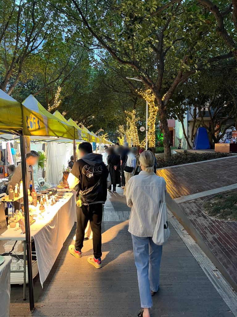 People waking in a Christmas Market with stalls selling ceramics at sunset when its starting to get dark. The trees in the background are lit up with lights.