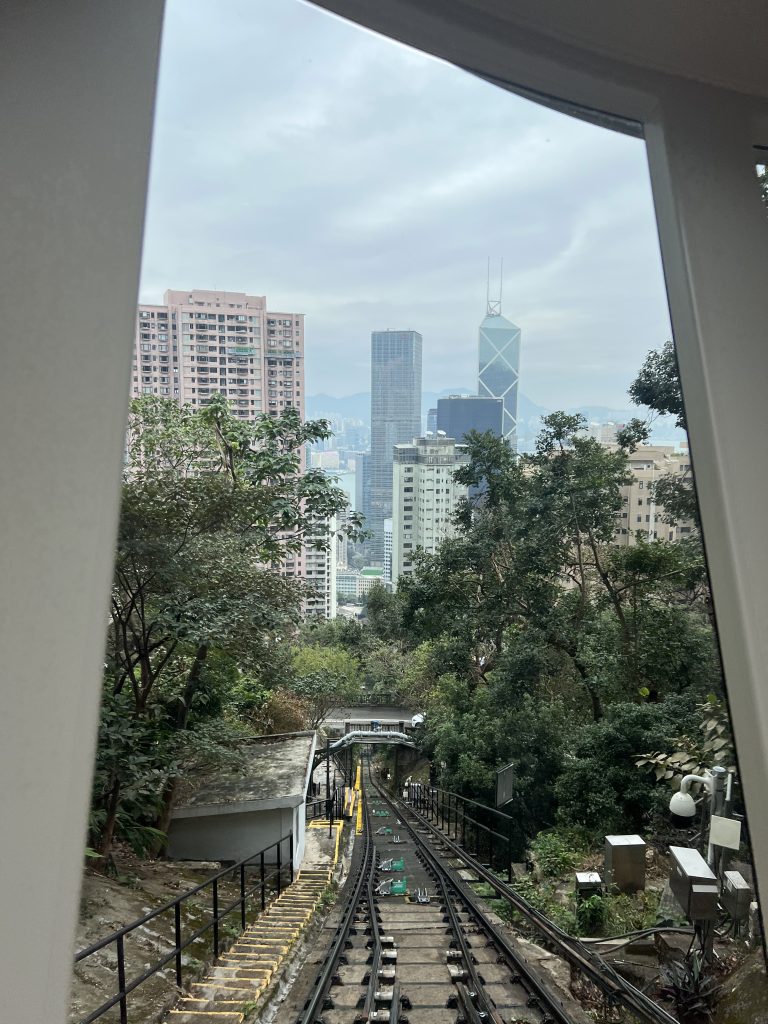 Train tracks leading downwards from the Peak Tram. The tracks are surrounded by forrest and in the background skyscrapers of Hong Kong skyline can be seen.