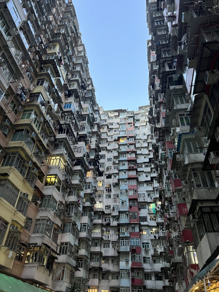 The Monster building of Hong Kong stretching into the sky. The building covers all sides, and has windows sticking out. The building is yellow, pink, blue and red in different vertical lines. 