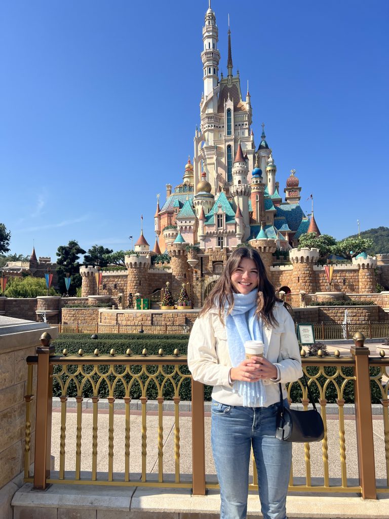 Girl standing in front of a miniature castle at Disney Land in Hong Kong. The castle is white with spires and caps in light blue.