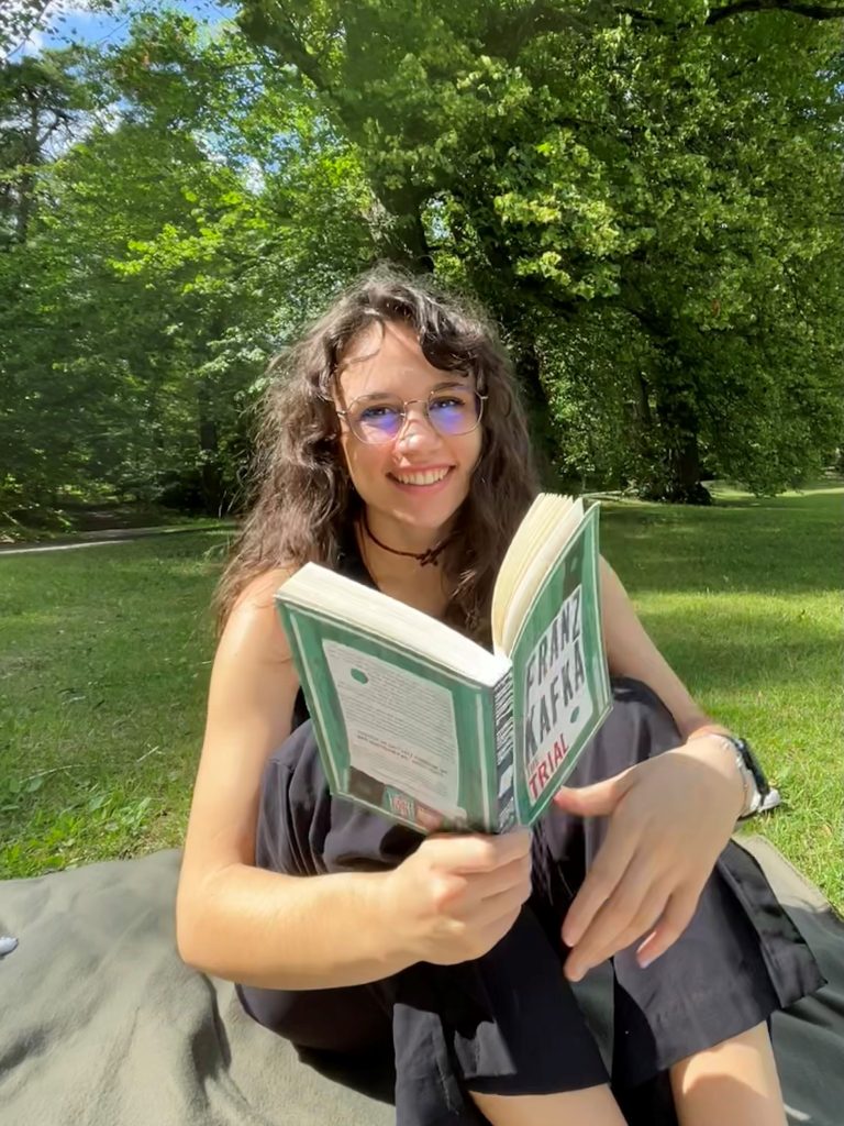 Kvinna med glasögon och mörkt hår sitter i en park ler och läser en bok.