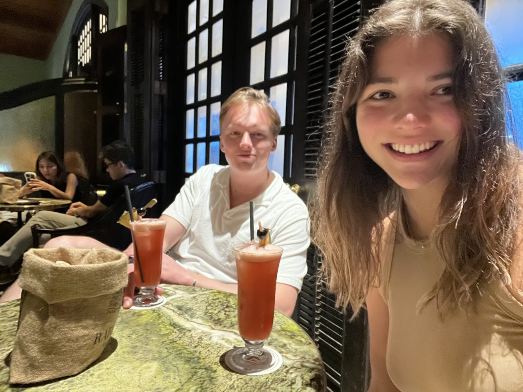 A boy and girl taking a selfie and smiling at the camera, sitting at a round table with two tall red drinks and a bag och peanuts.