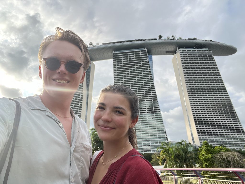 A couple taking a selfie infront of Marina Bay Sands, a hotel with three towers and what looks like a long boat stretching on top of the three towers. 