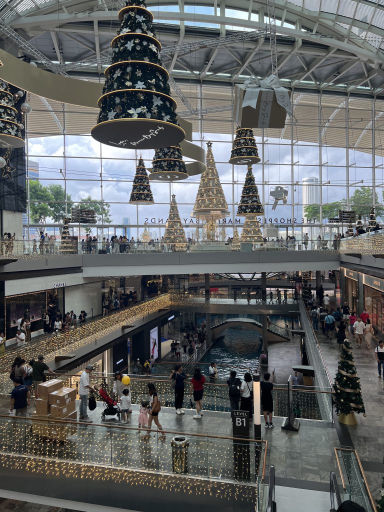 Inside of Shoppes by Marina Bay where you can see a channel cutting through the room, two floors with glass windows in the background. Christmas trees and presents are hanging down from the ceiling and people are walking around with shopping bags.