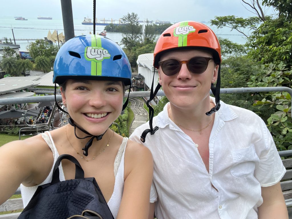 A couple in colorfull helmets sitting on a skii lift taking a selfie. In the background you can see greenery, the ocean and several large ships. 