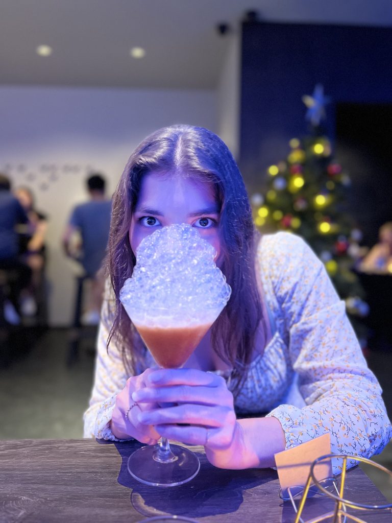 A girl looking out over a drink with bubbles like a bubble bath. The background is blurry, where people can be spotted and a christmas tree.