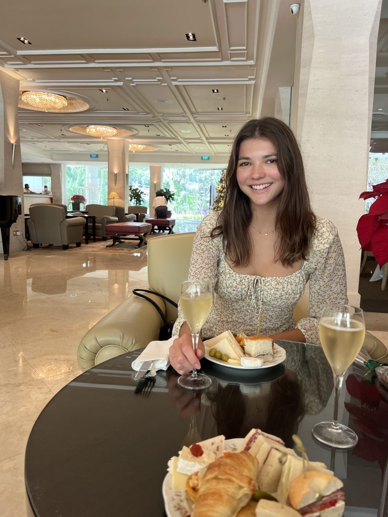 Girl dressed upp in a yellow flowery dress sitting at a round table with sandwiches and a glas of champange, with a hotel lobby behind her, filled with little soffas and glas doors.