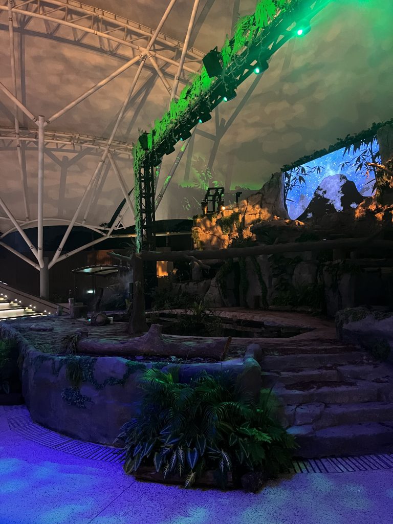 A stage with a pond and a wooden beam going over the stage. The ceiling is white and there are steps leading up to the stage. There is a projector showing the night sky and behind the stage, seats can be spotted.