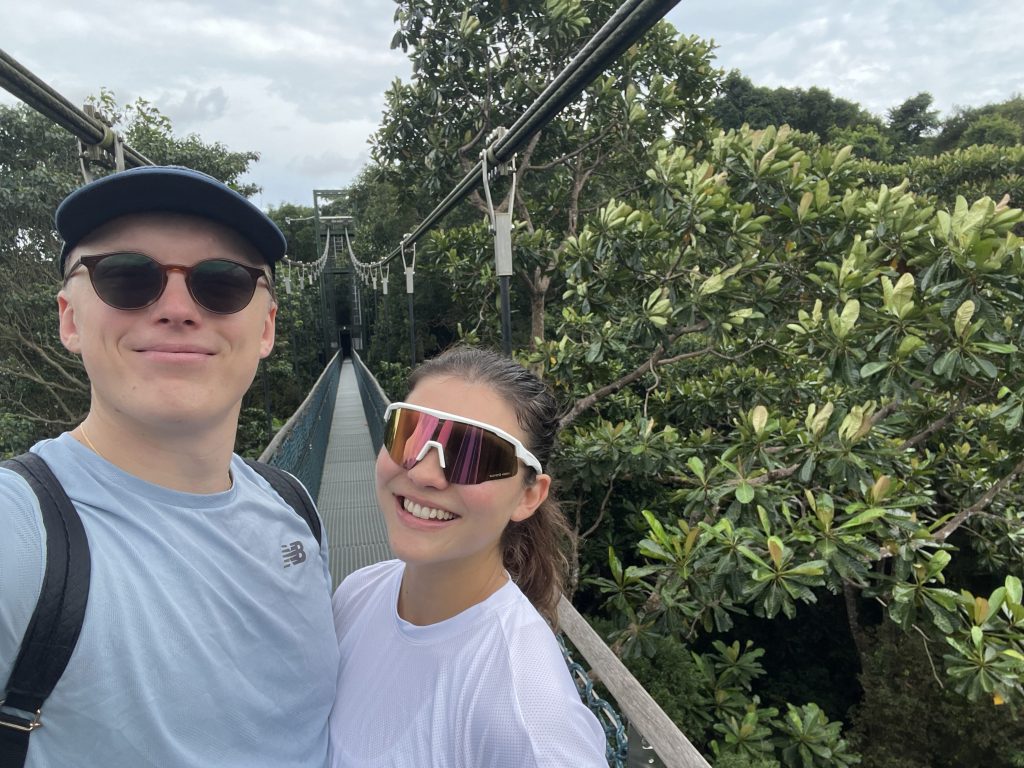 A girl and boy in training gear on a suspension bridge taking a selfie. They are surrounded by tree tops and green leaves. 