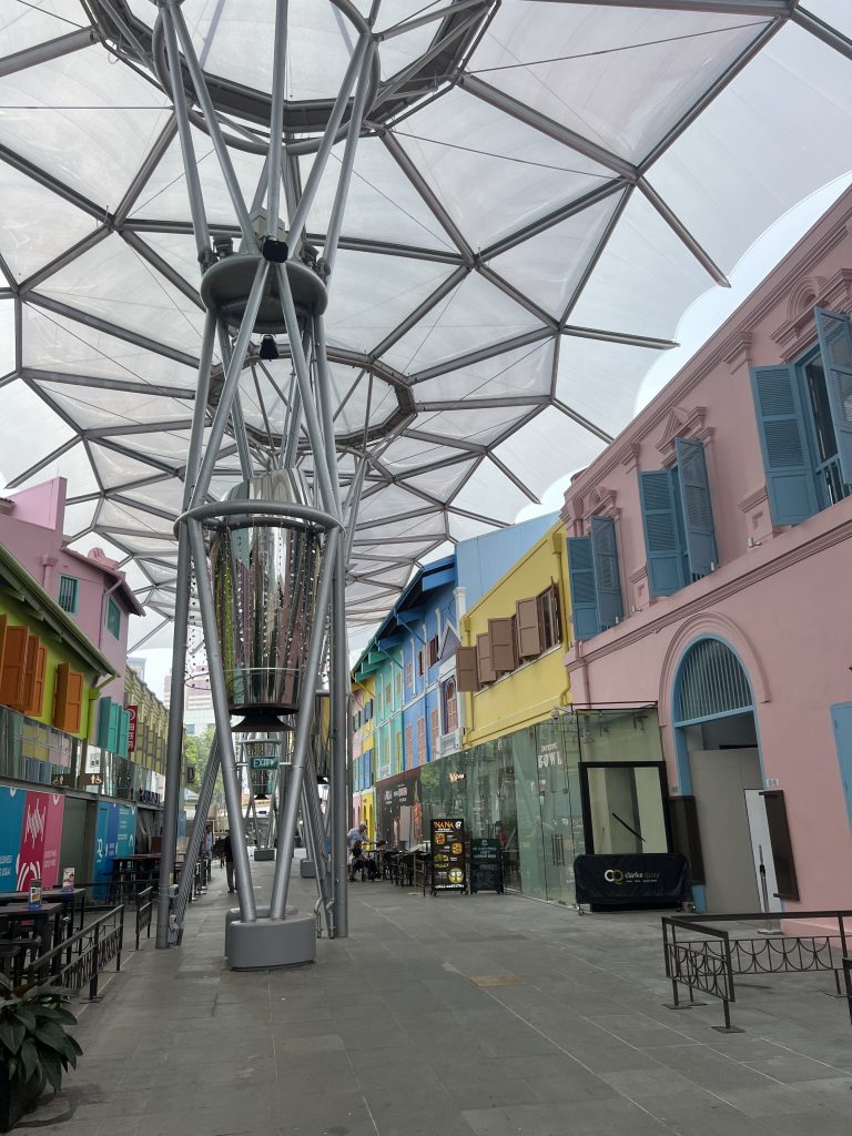 A road with colourful houses including a pink house with blue window shades, a yellow house with brown window shades and a blue house. From the road, giant metal structures stretch into the air like giant umbrellas.