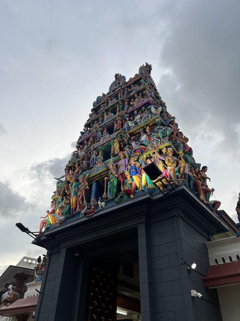An entrance to a hinduistic temple. the gate is black and square and on top, shaped like a pyramid are levels of several statues in bright colors. The statues are of human form mainly, although some cows can also be spotted.