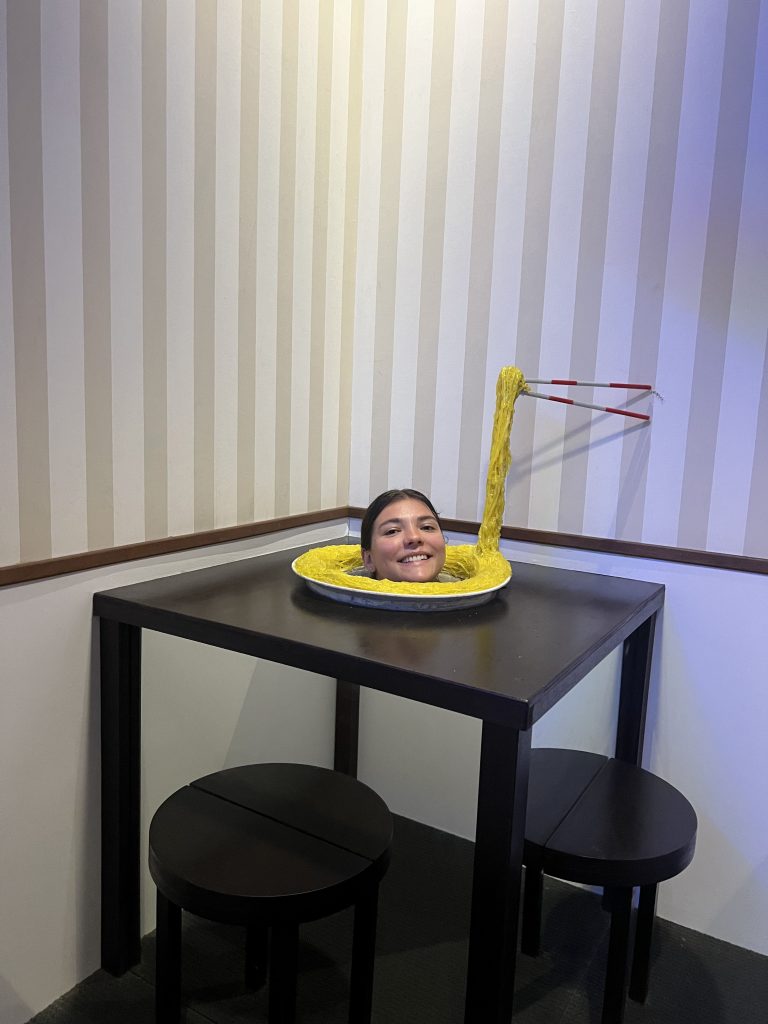 A girl peeking out with her head of an optical illusion making it look like her head is in the middle of a plate of spagetti on top of a table with chairs.