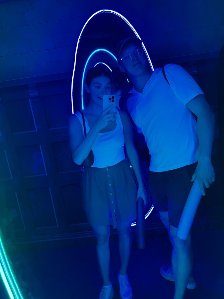 A girl and boy taking a selfie in a mirror. The light is blue. They are holding a pool noodle each.