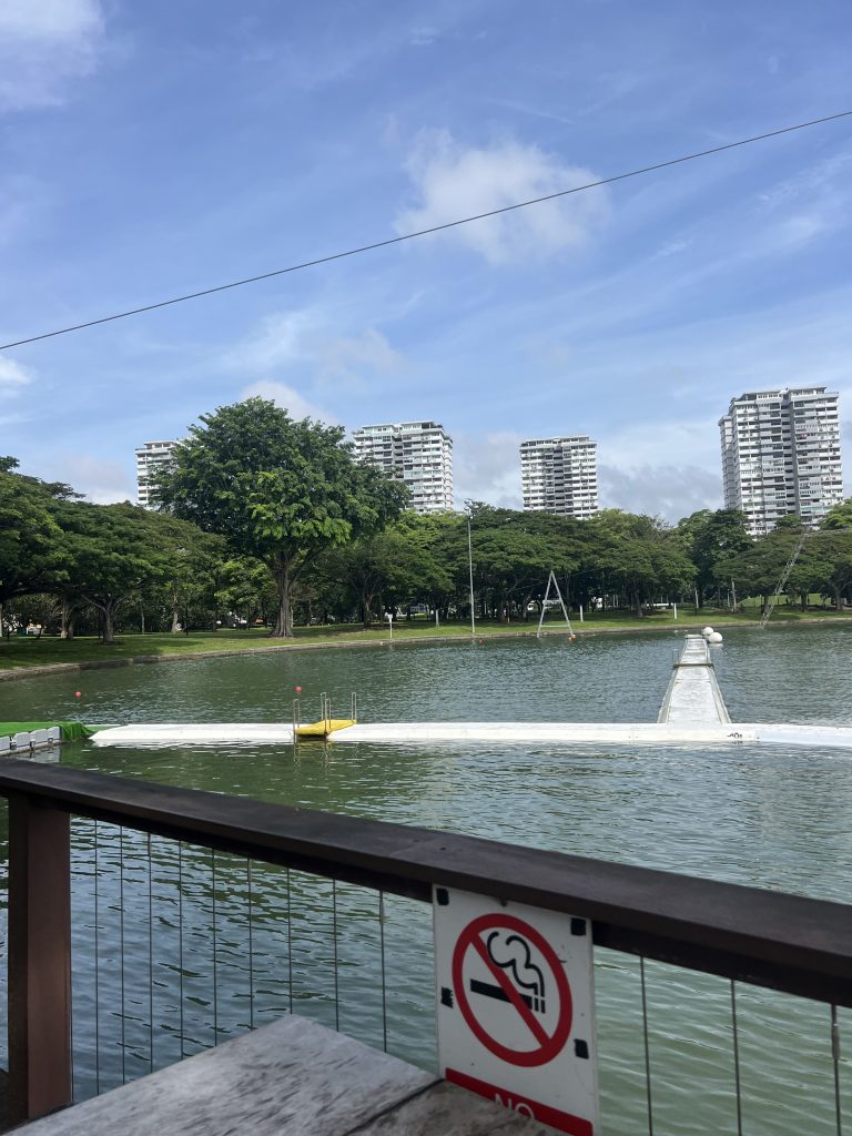 A pond with a long white buoys that you could walk along. In front is a fence with a "no smoking" sign in an the background is a garden with trees and three tall buildings.