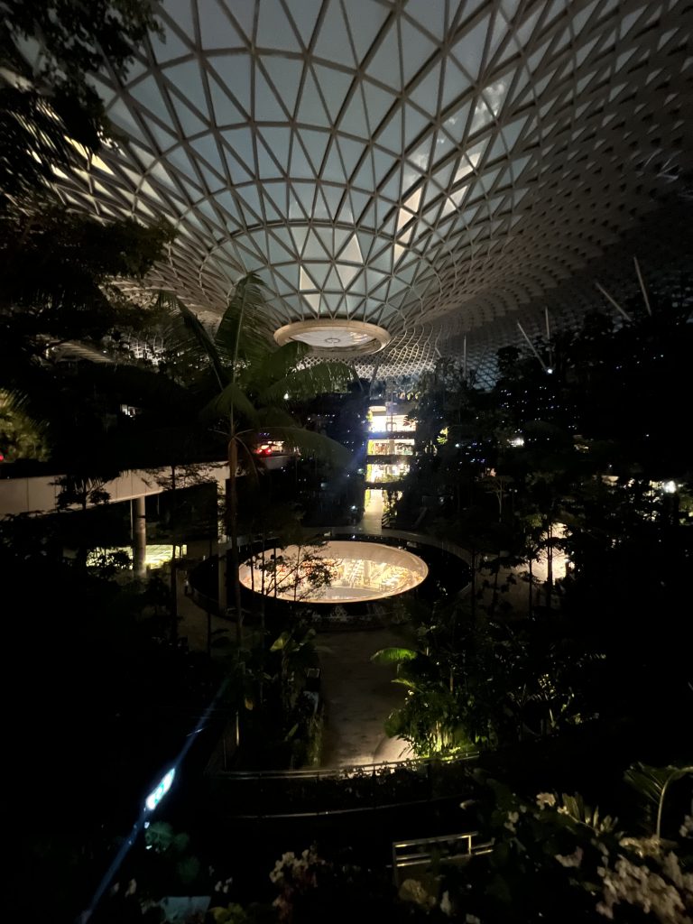A dark large room with a glass dome stretching downwards in the ceiling. At the bottom a lit up waterfall can be seen and it is surrounded by plants.