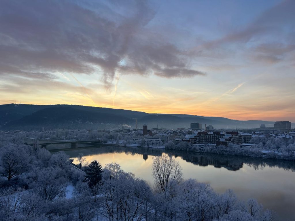 View of a sunrise above Heidelberg. The photo shows the orange sky, trees and buildings covered in snow and ice and the river reflecting the image of the sky. There is a bridge over the river.