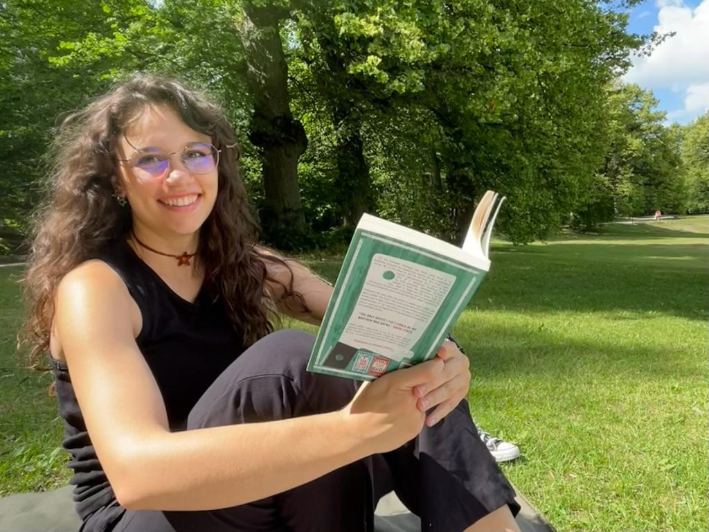 Photo of a young woman smiling. She is wearing a black tank top and black pants and has her hair down. She is holding a green book and sitting on a blanket. In the background there is a park with green grass and trees.