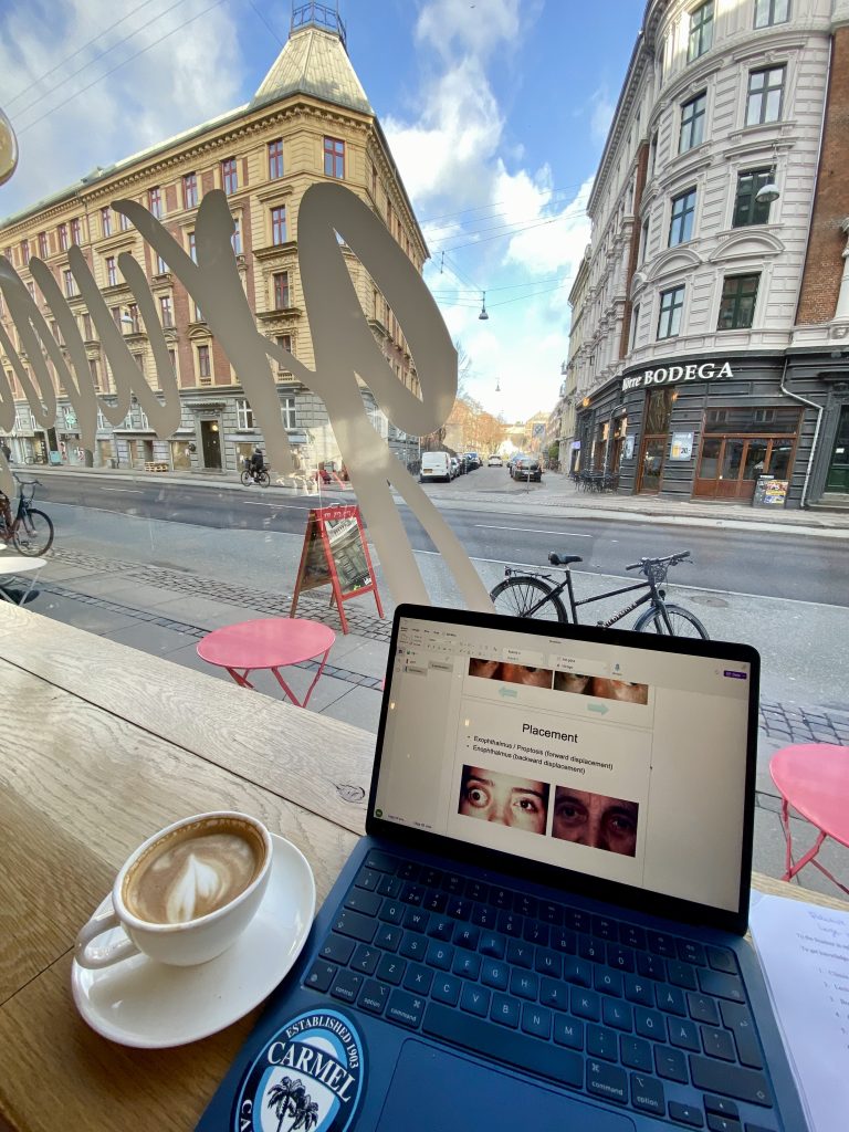 Dator med föreläsning i oftalmologi bredvid en cappuccino, som står på ett fönsterbord med utsikt över en gata