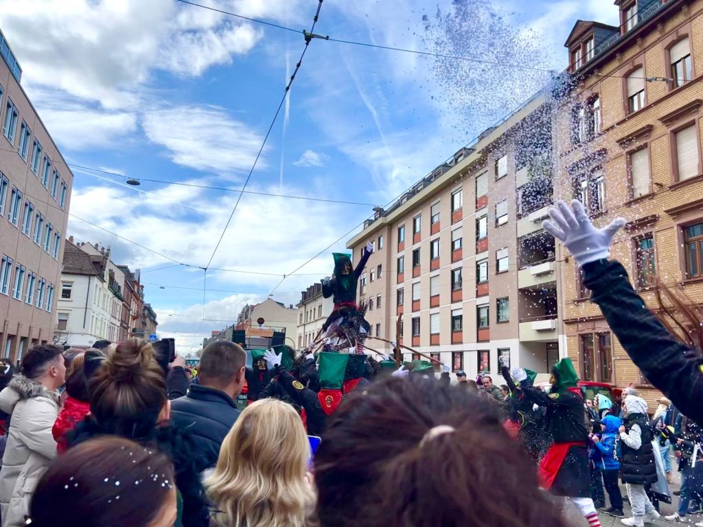 Performer in the parade being lifted up by his team. The person is wearing black and green clothing and a large wooden mask of a caricature face. On their head they have a green hat and a wig. At the front of the photo the crowd can be seen and on the right a hand is throwing confetti in the air. 