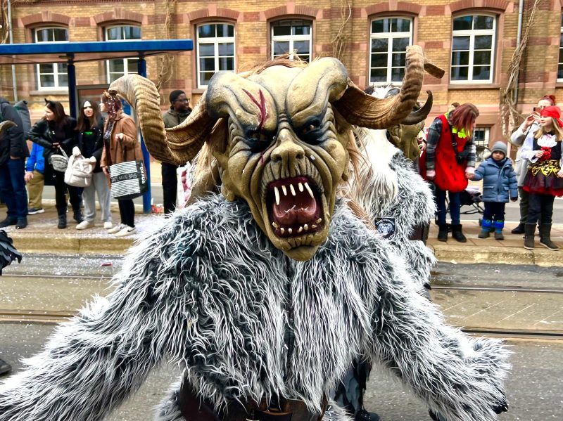Person wearing a large scary wooden mask. They have twisted horns at the side of the head and the mask is a beige face with an open mouth. The mask has pointy white teeth and a big tongue. The eyes are dark and there are scars painted on the mask. The person is wearing a furry gray suit and in the background a crowd of people and an apartment building can be seen.