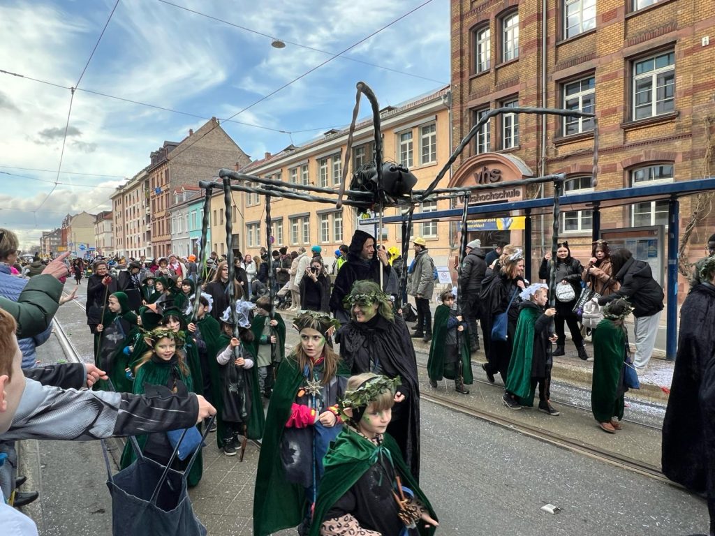 Performers in the parade dressed up in black and wearing elf ears and leaf crowns. A giant spider sculpture is held up above them. They are walking down a street and in the background the viewers and apartment buildings can be seen. 