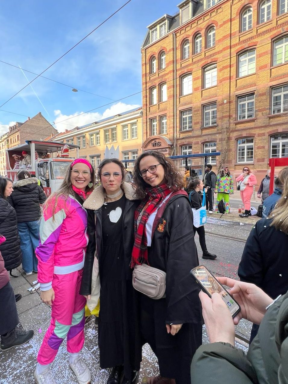 A girl dressed in a neon pink track suit, a girl dressed in a black dress and wearing a crown and queen like make up and a girl dresses as a wizard all posing for the photo. I the background there is a parade, a crowd of viewers and apartment buildings.