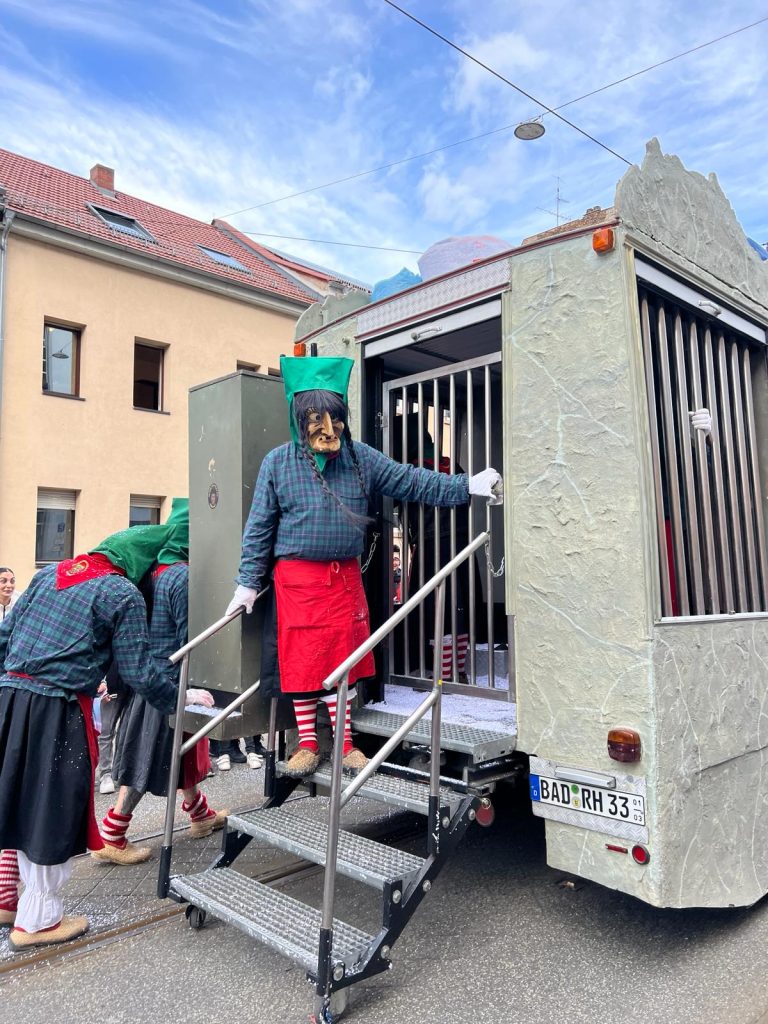 A float in the parade with a installation representing a prison cell. A performer is standing on the back of the float on some stairs. They are wearing a red apron and a dark blue shirt and have a caricature wooden mask on their face. They are wearing a green hat and a wig with two braids.