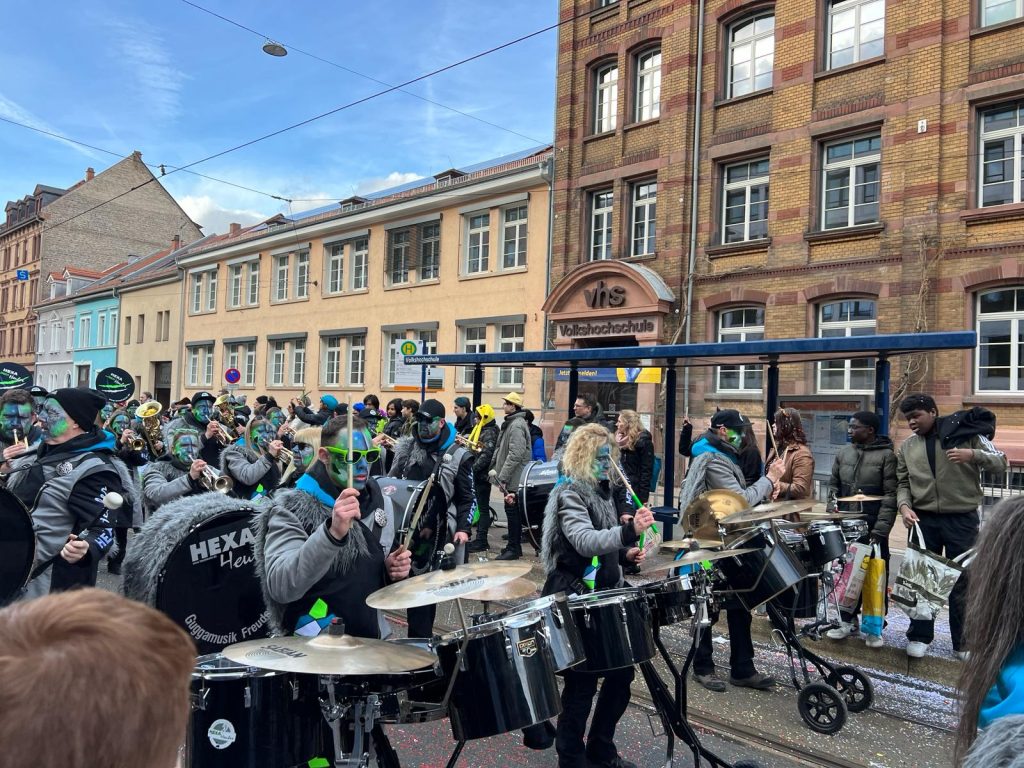 A marching band walking down the street. The performers in the photo are playing instruments such as drums and have their faces painted with a green and blue scenery. Apartment buildings can be seen in the background.