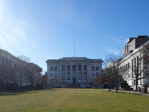 The white and gray marble building of Harvard Medical School in the sun behind a small grassy area.